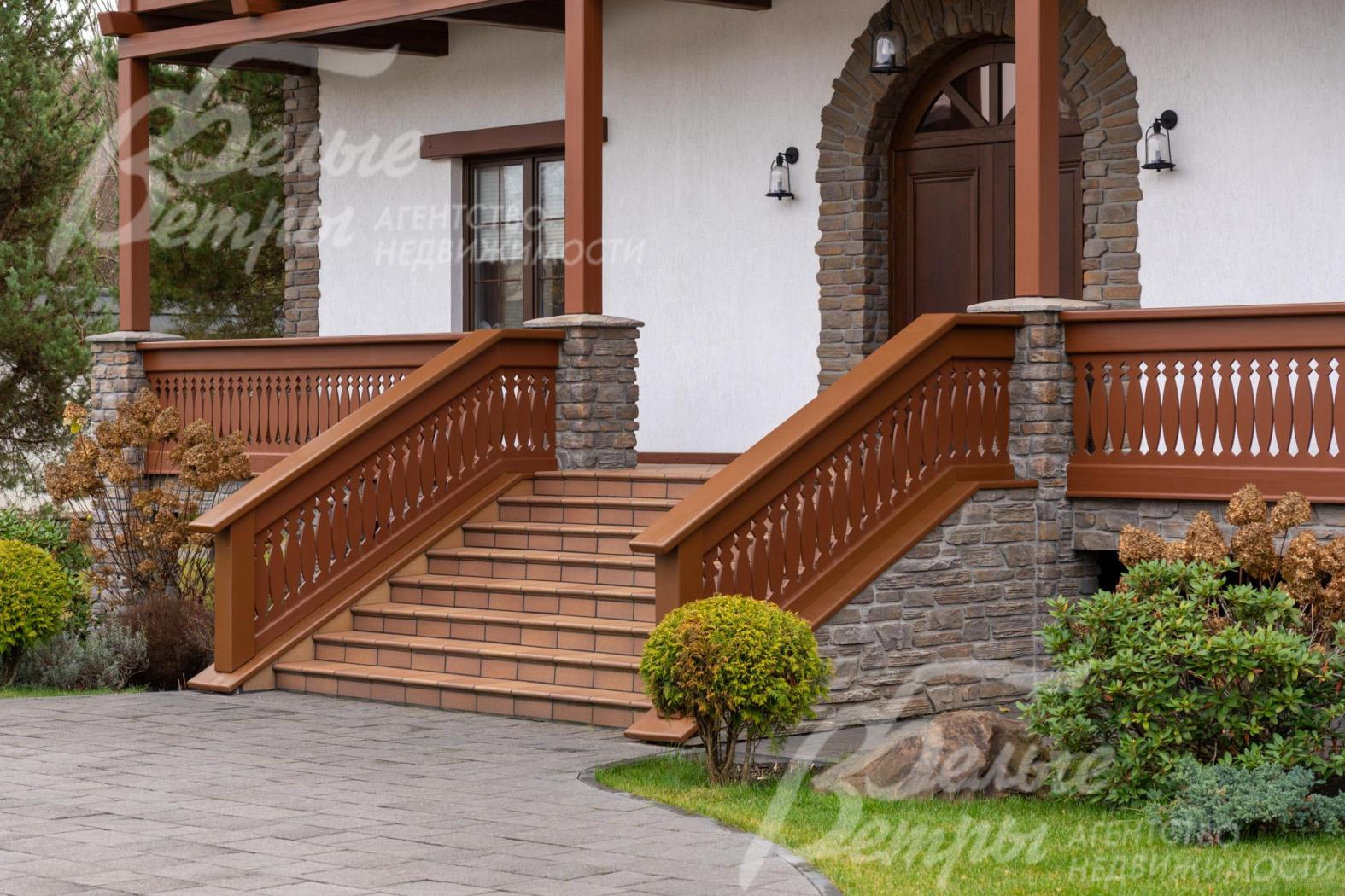 Главный вход в дом эффектно декорирован искусственным камнем. К крыльцу дома поднимается лестница из клинкерной плитки тёплого терракотового оттенка с деревянными перилами с резными балясинам